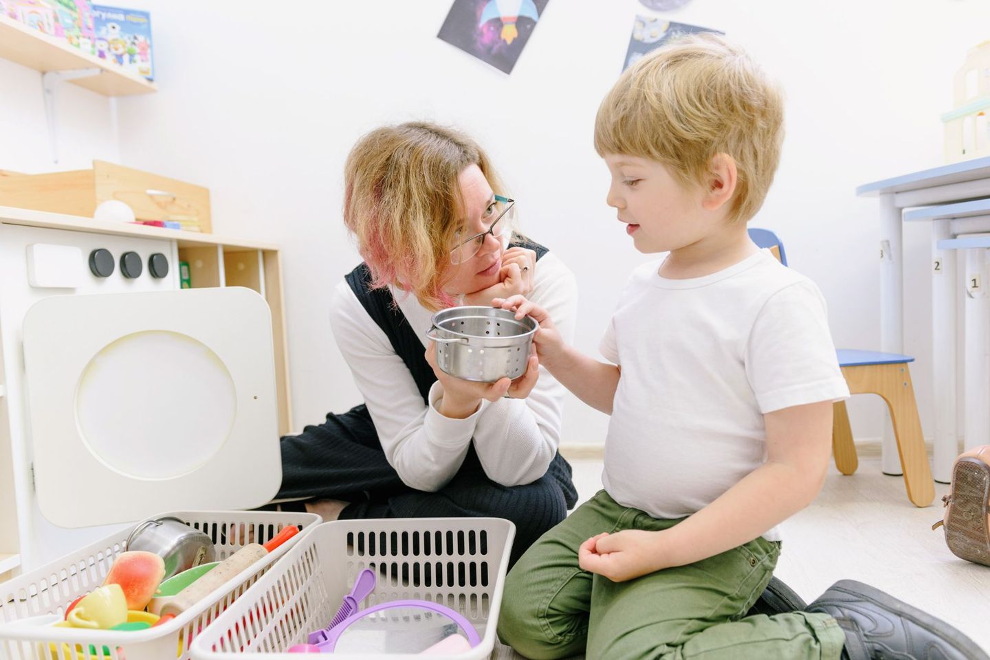 A clinician sitting on the floor of a therapy room, listening closely to a young boy holding a small steel pot
